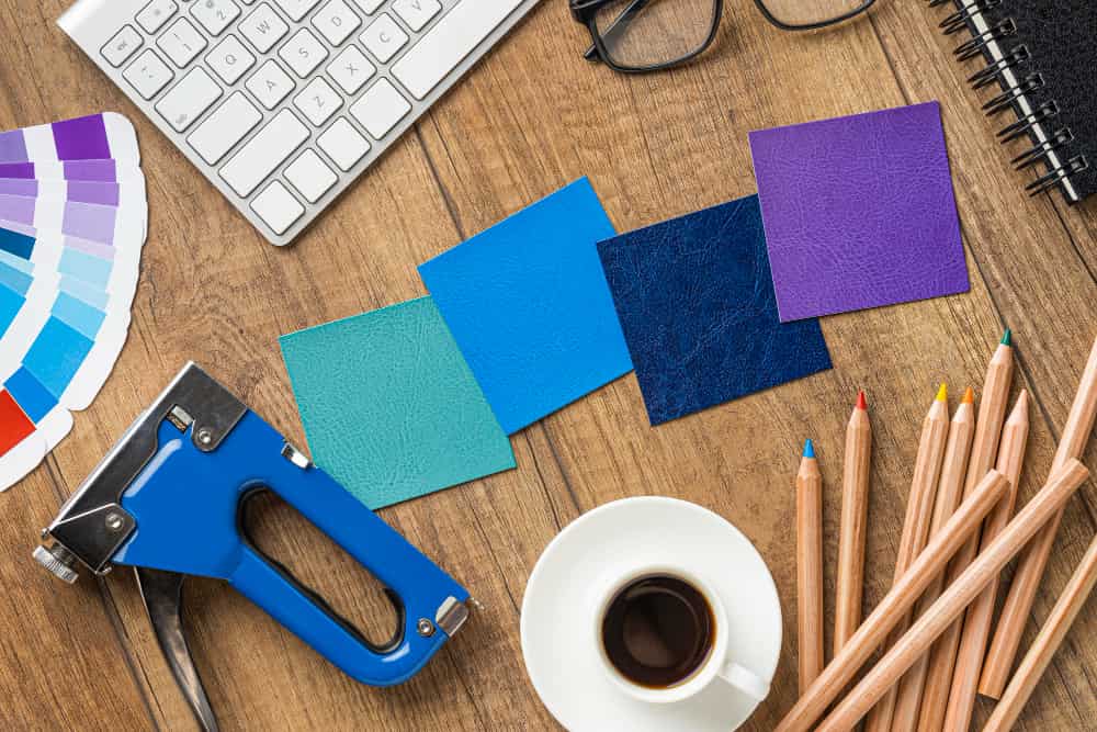 un poinçon, des morceaux de cuirs, des crayons de couleurs, un nuancier, posés à côté d'un ordinateur