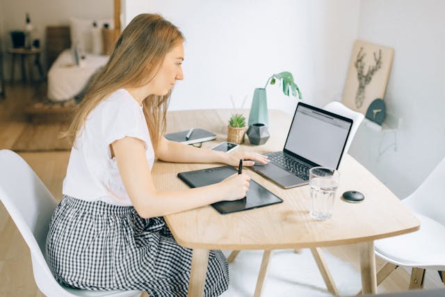 une femme qui travaille sur son ordinateur posé sur une table avec des plantes vertes et un verre d'eau, agence digitale vs freelance