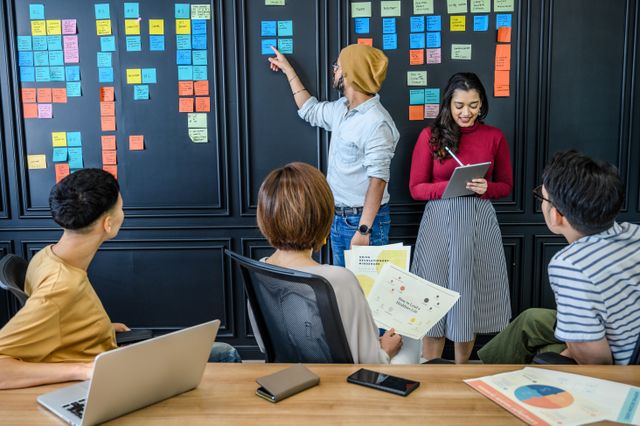 une homme en hemise bleu qui pointe un post it du doigt sur un mur noir plein de post-is devant une équipe, agile transformation